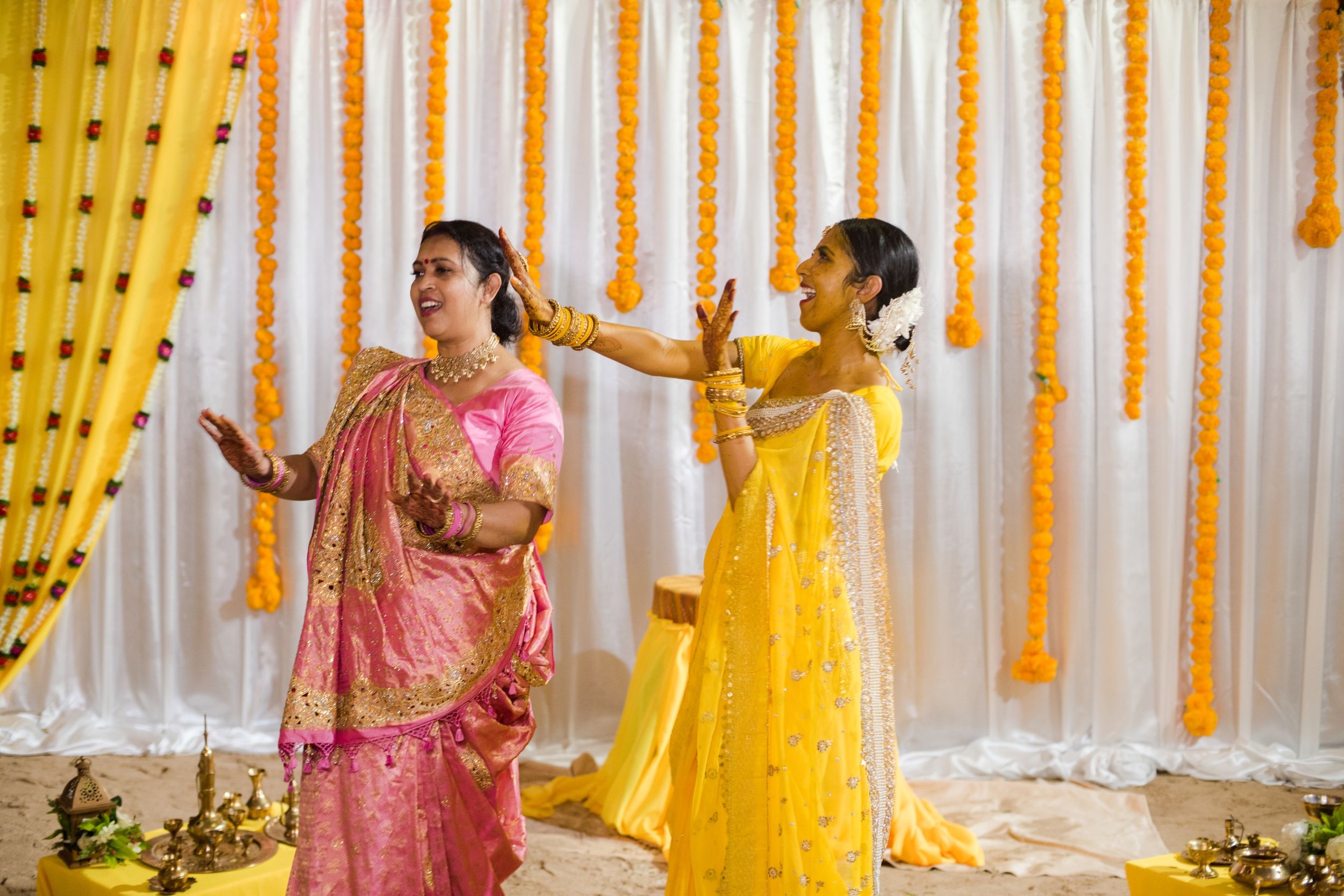 Two women in colorful traditional attire performing dance moves against marigold decorated backdrop.
