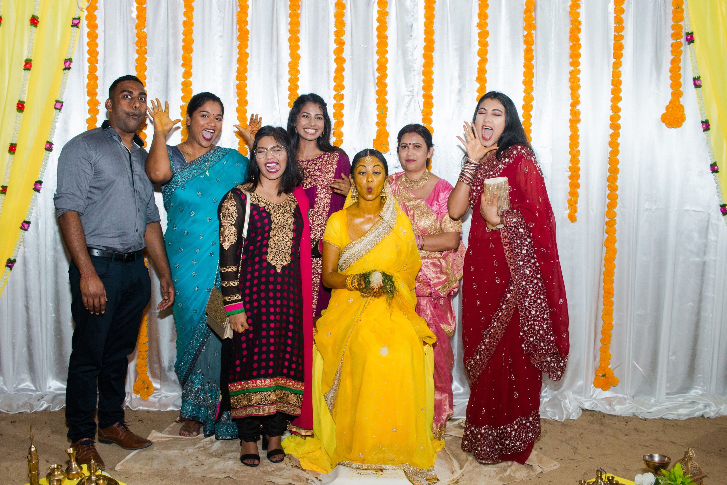 Group photo of wedding guests in traditional Indian attire against floral decorated backdrop.