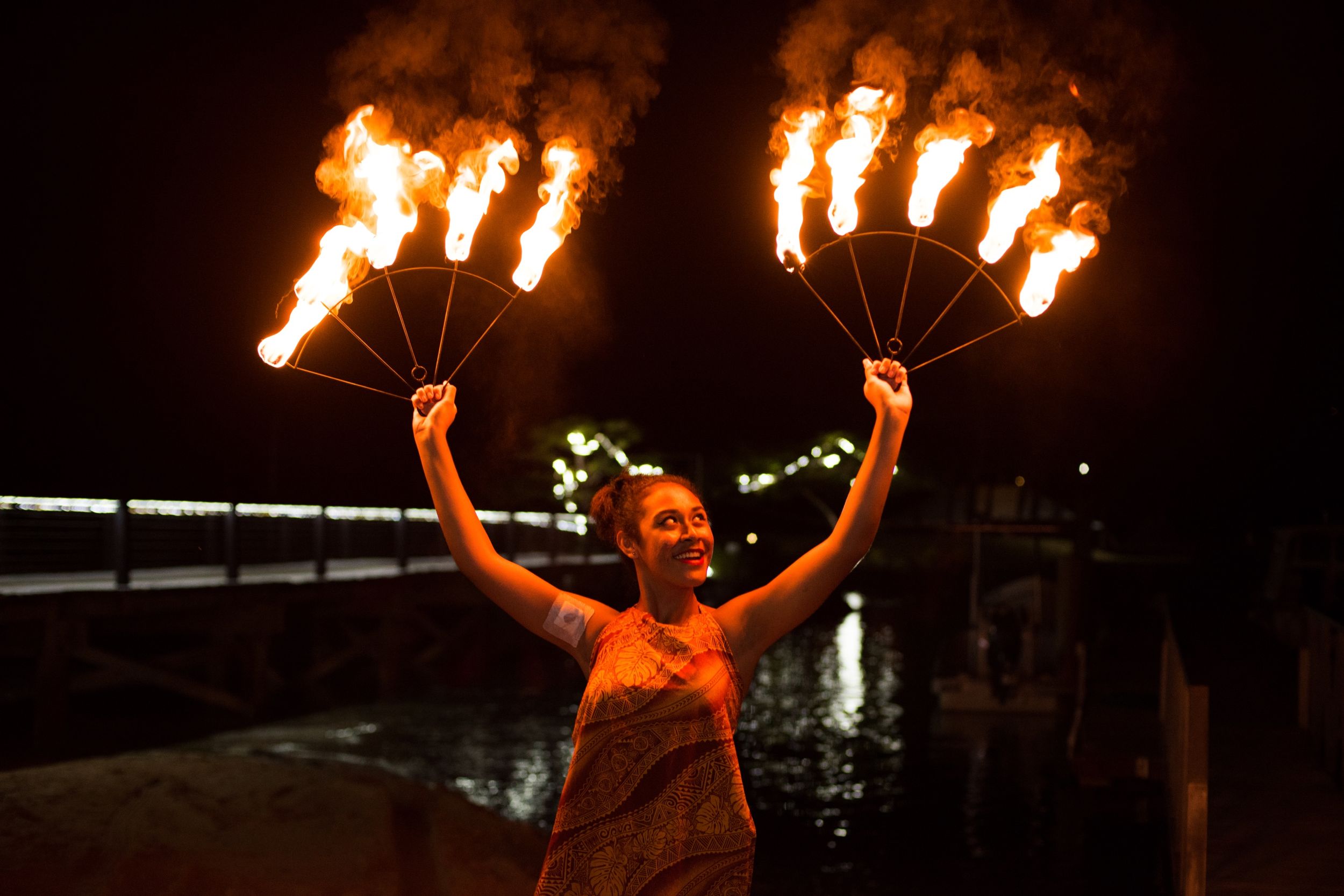 Fire performer holding up two flaming fans at night near water.