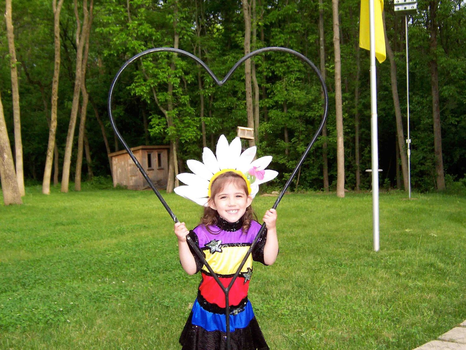 Child wearing colorful dress and daisy crown standing under heart-shaped arch in garden setting.