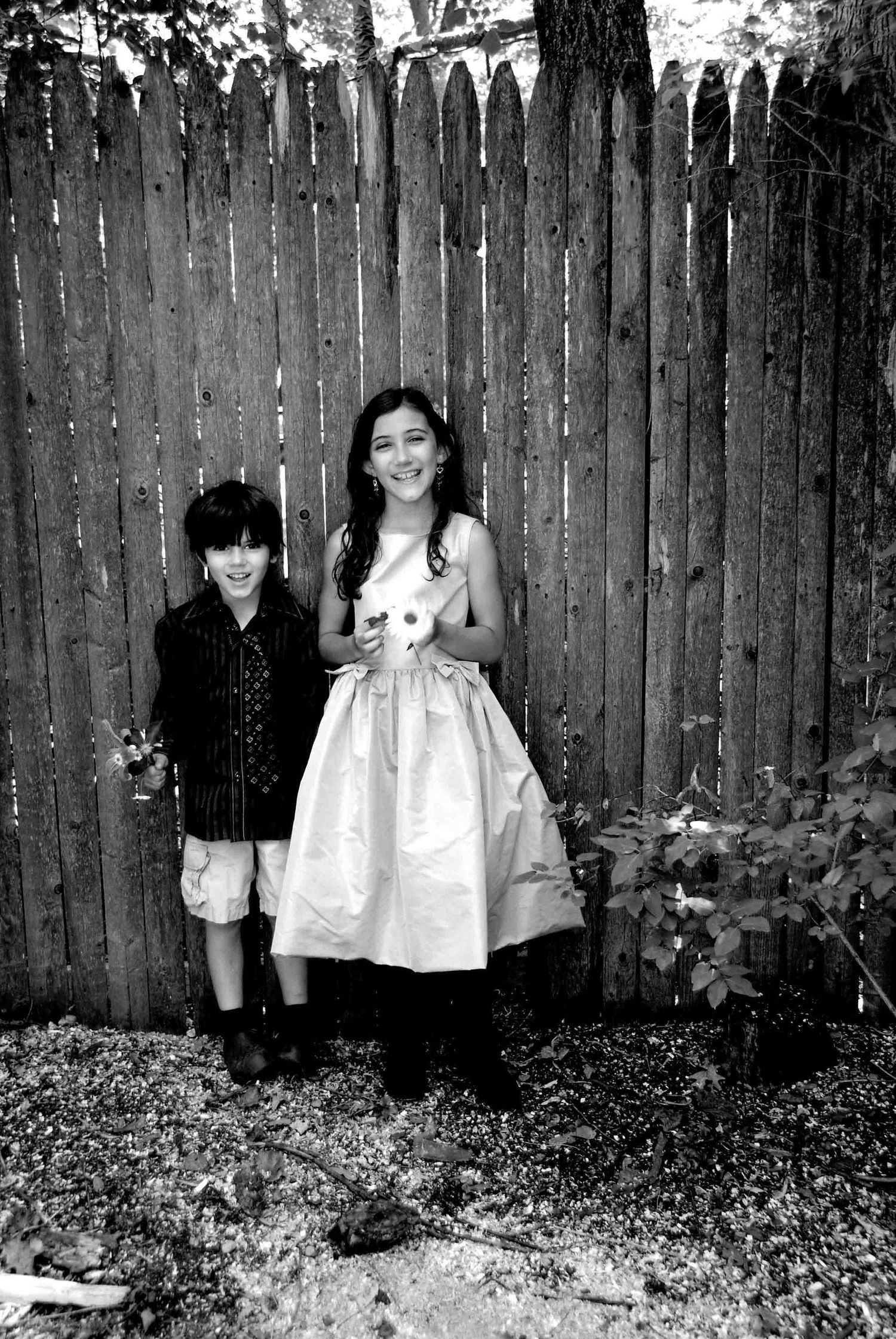 Black and white portrait of two children standing against wooden fence backdrop.