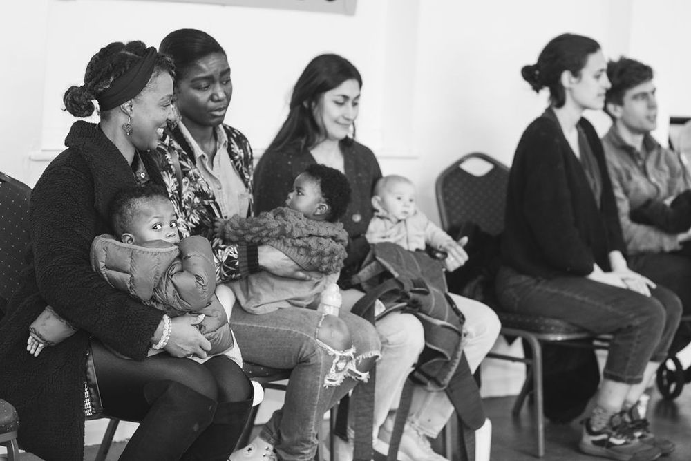 Parents and babies sit together in chairs during what appears to be a group gathering or meeting.