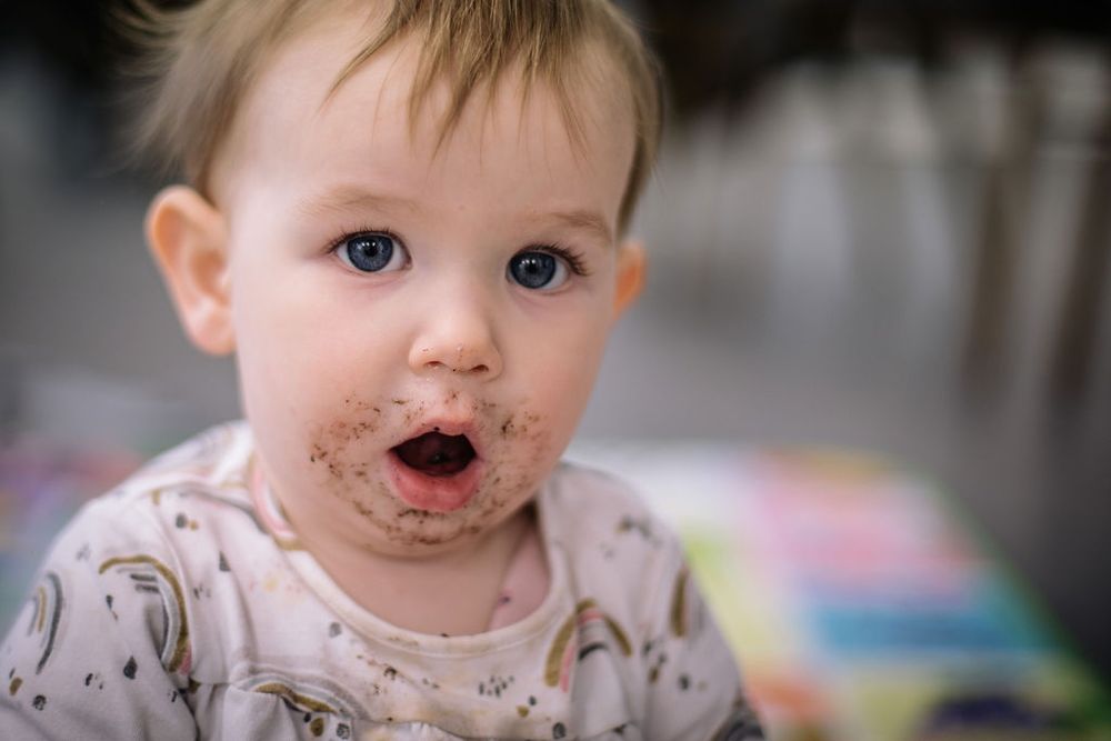 A toddler with food on their face wearing a rainbow print shirt looks at the camera with surprise.