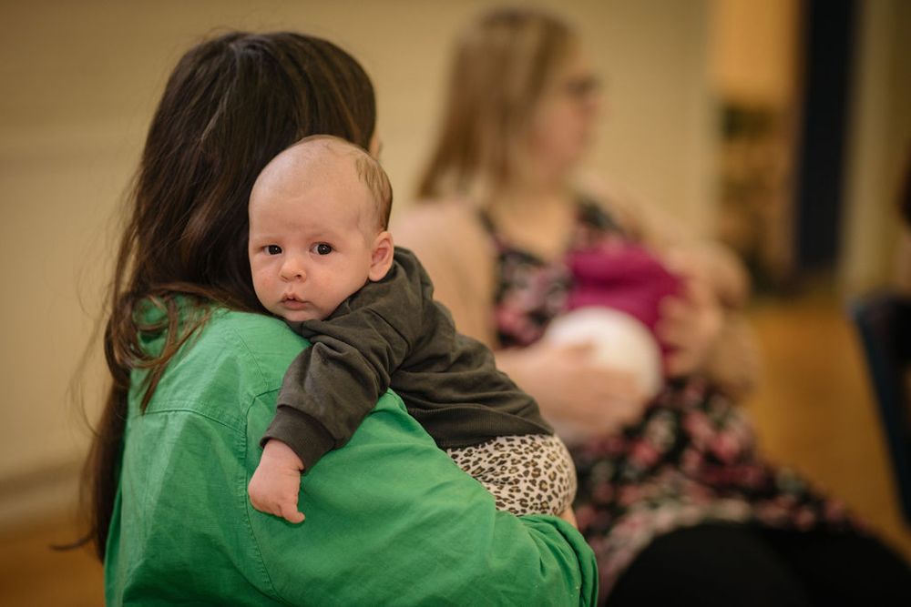 A baby in gray clothing being held while others gather in the background of a community space.