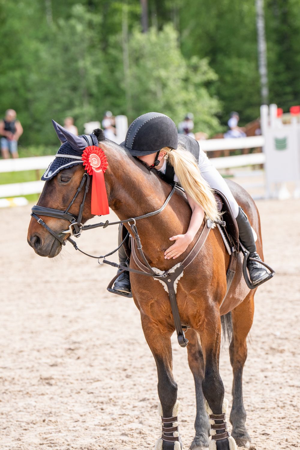 Equestrian rider affectionately hugs brown horse wearing red ribbon during competition in outdoor arena.