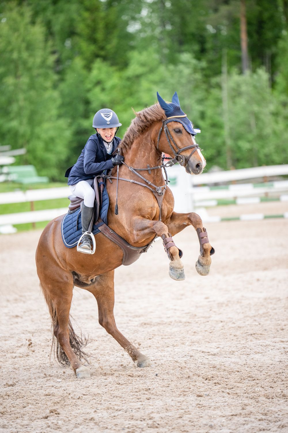 Bay horse and rider clearing a jump during equestrian competition on sandy arena.