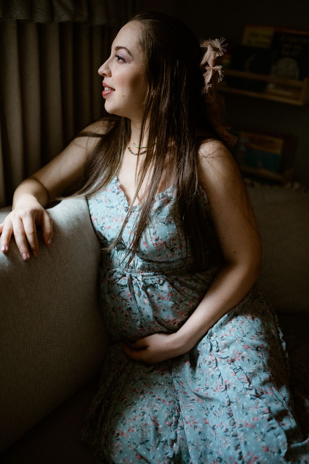 Side view of pregnant woman in blue floral dress lounging on couch in moody lighting.