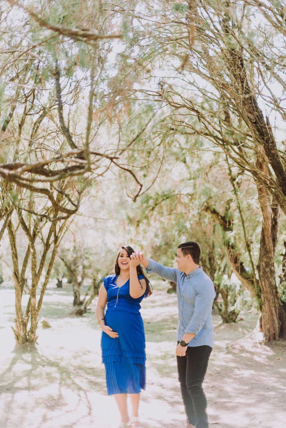 A couple shares a joyful moment outdoors among trees with her in a flowing blue dress.