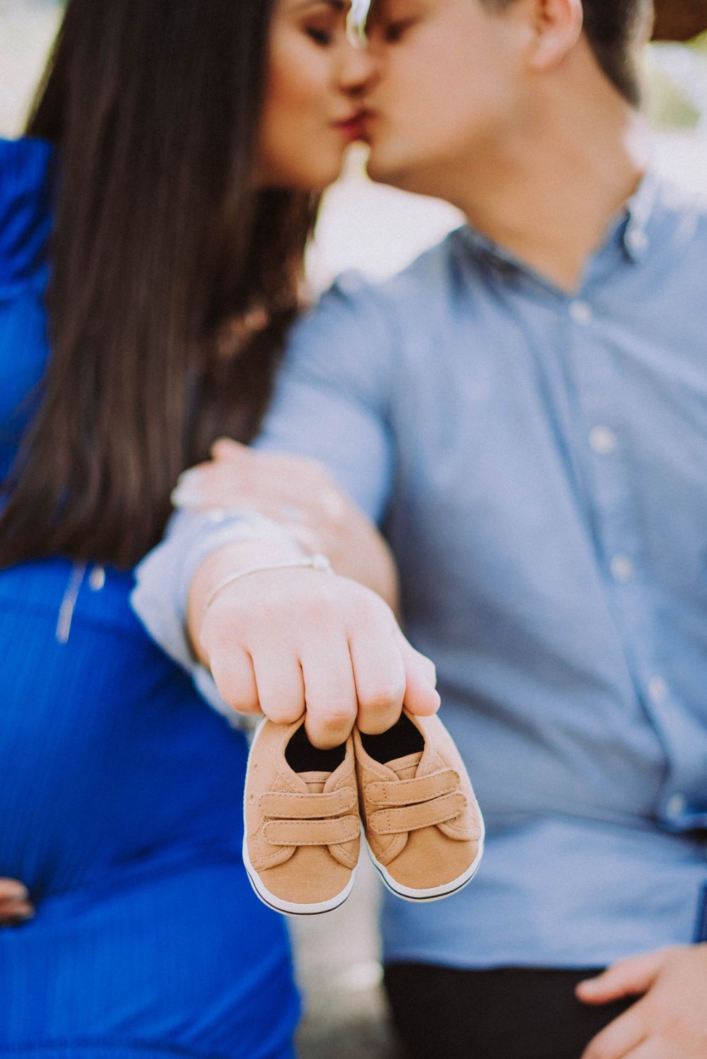 A couple kisses while holding tiny beige baby shoes in the foreground.