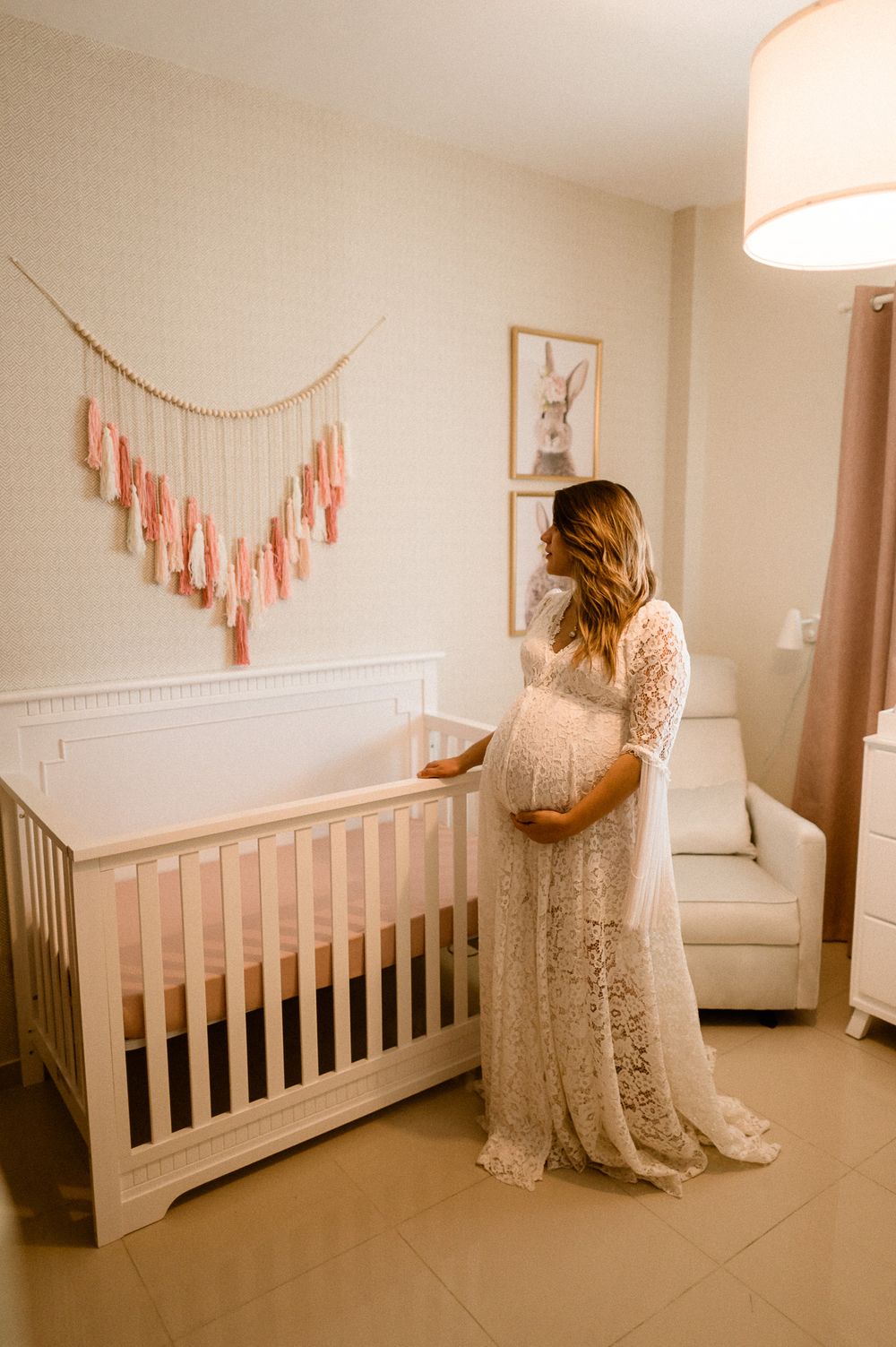 Maternity photoshoot in nursery with white crib and pink tassel wall hanging decoration.