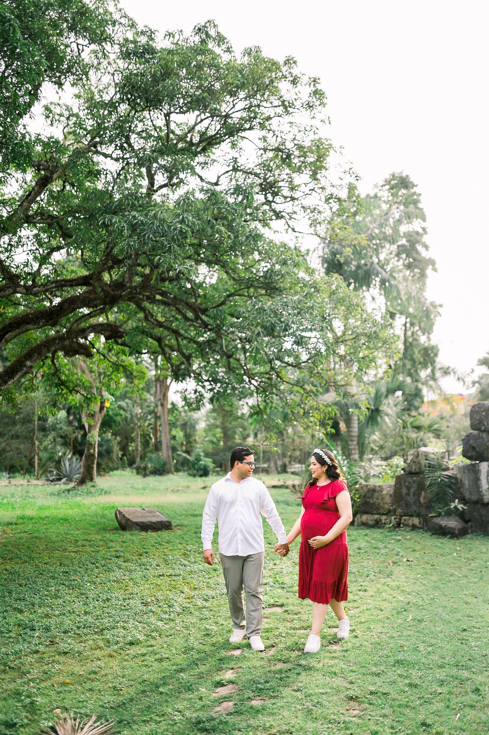 A couple takes a romantic stroll through a lush green garden path under towering trees.