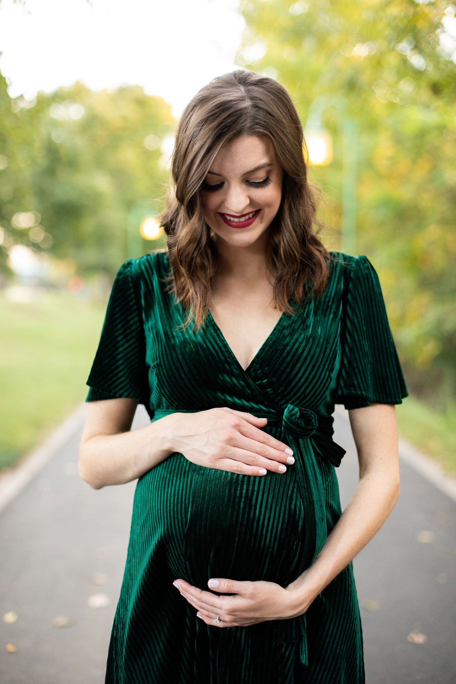 Woman in emerald velvet dress tenderly touches her baby bump in outdoor maternity photos.