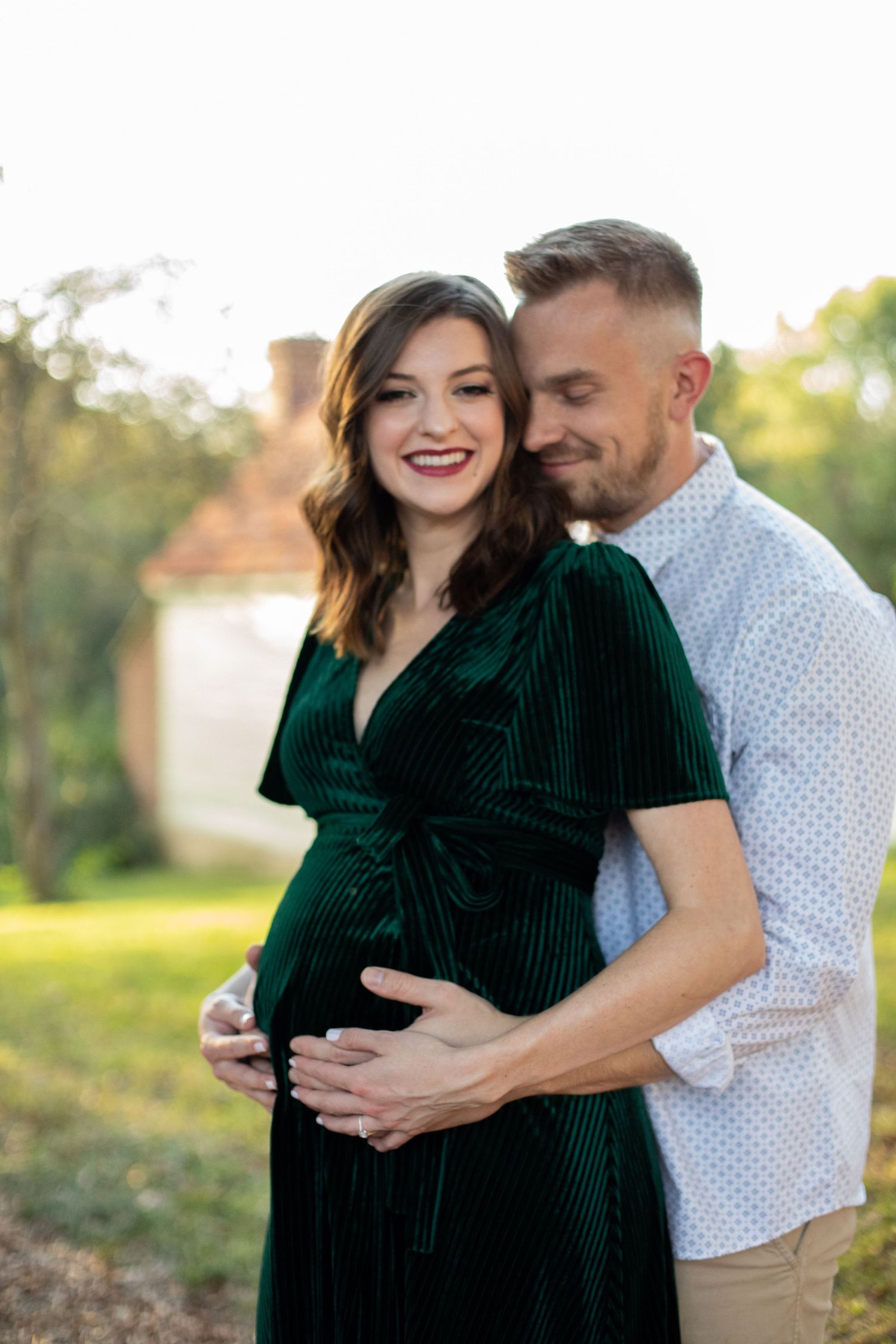 A couple embraces outdoors during a maternity photoshoot with golden sunlight in the background.