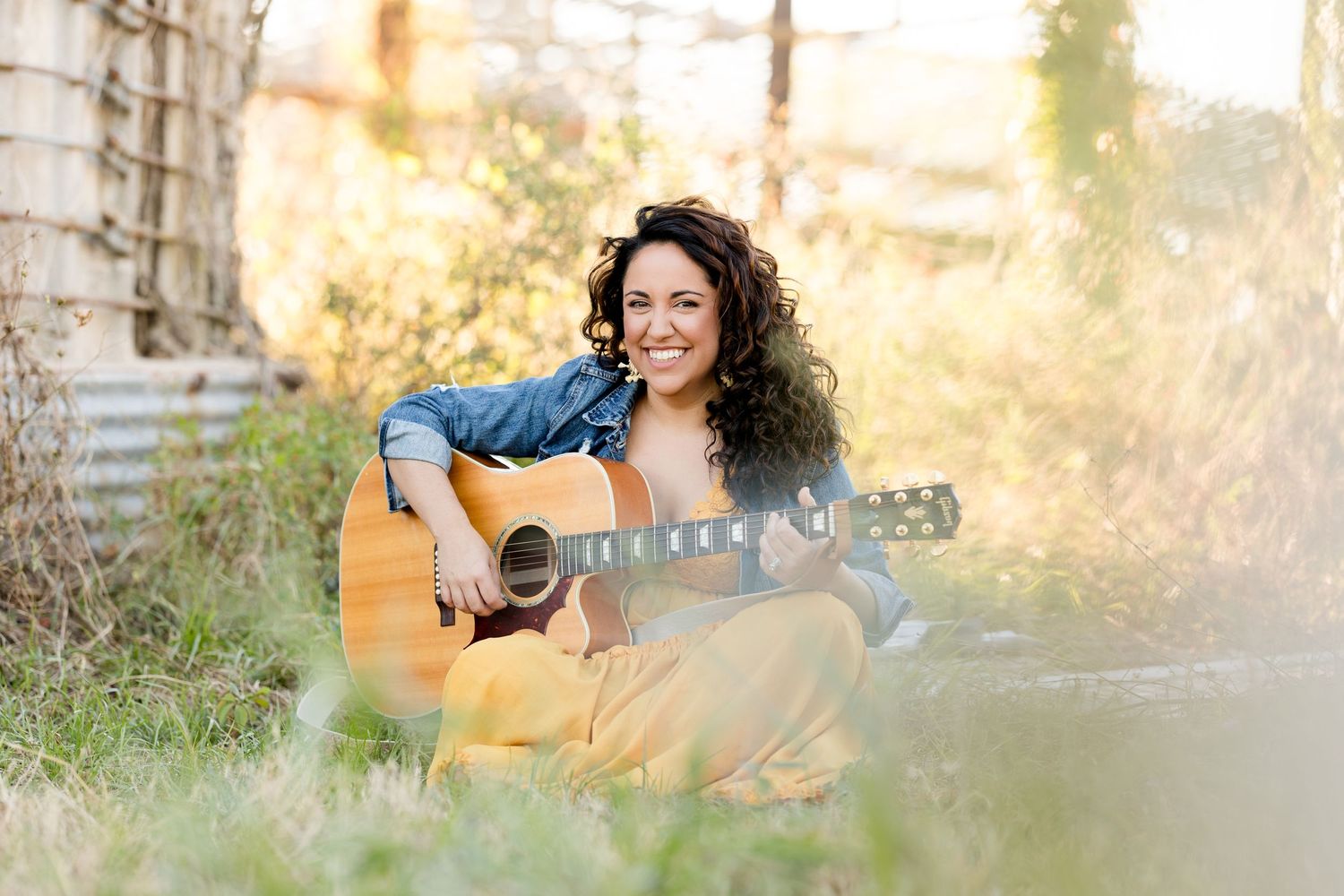 Woman poses casually in field during sunset with warm golden lighting.