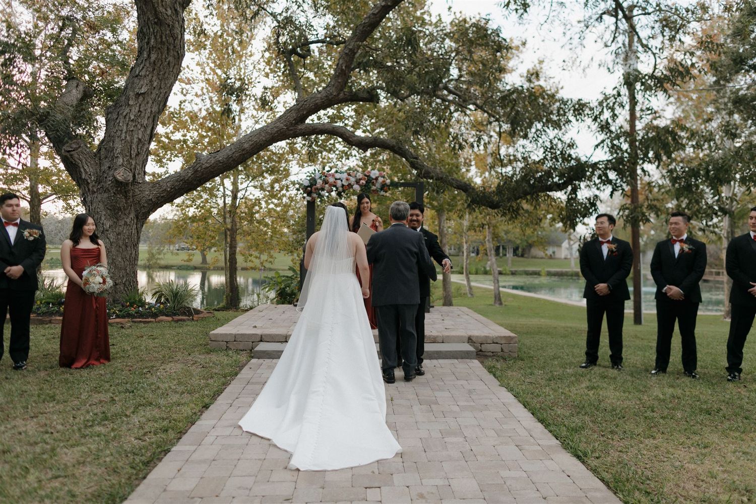 A wedding ceremony under large oak trees with guests standing alongside a stone pathway.