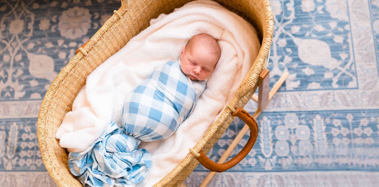 Sleeping baby in blue gingham outfit rests peacefully in woven bassinet on patterned rug.