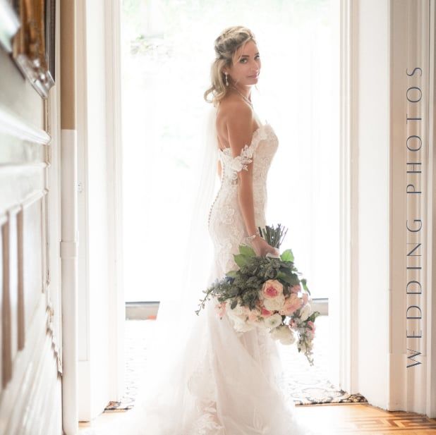 A bride in lace gown poses in doorway holding a romantic bouquet with greenery.