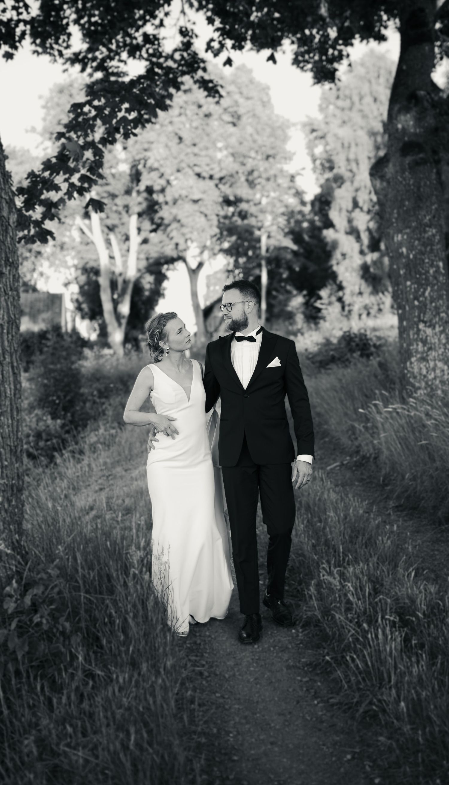 Black and white wedding portrait of a couple walking along a tree-lined path.