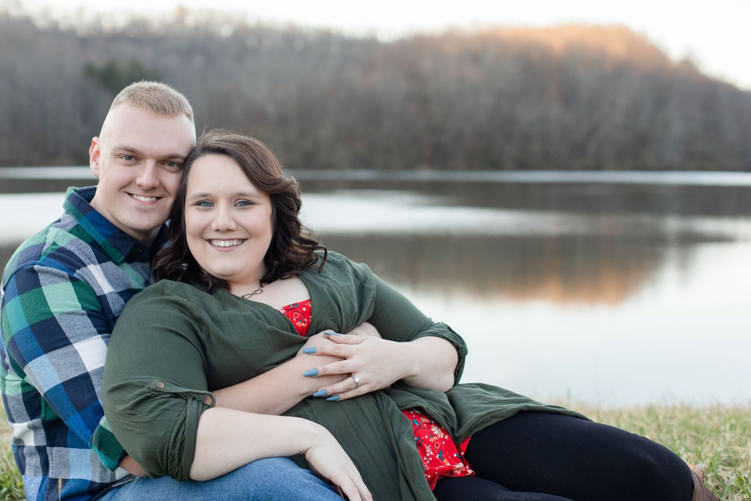 A couple embraces by a serene lake during an outdoor photoshoot at sunset.