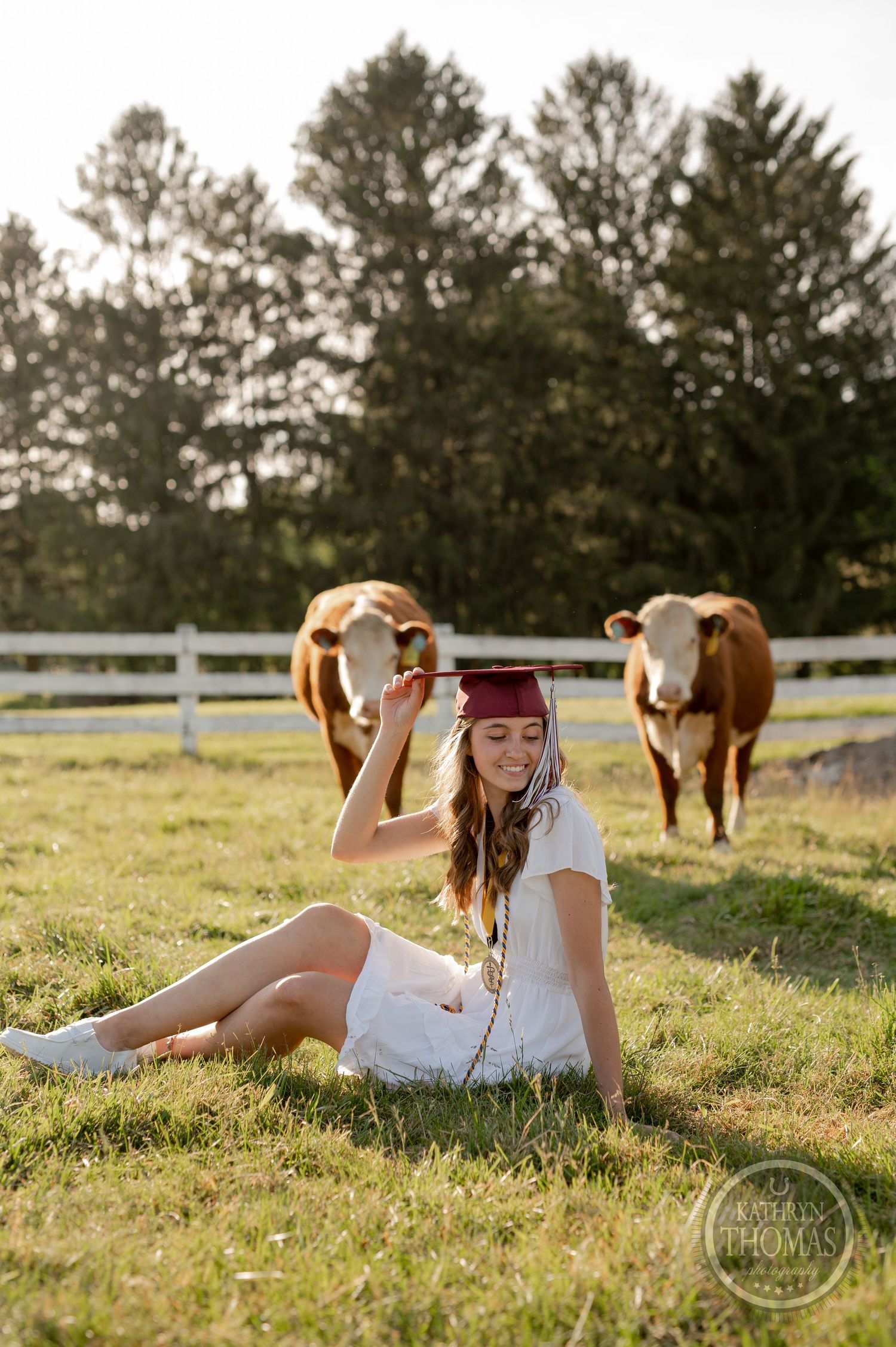 Graduate in white dress and red cap poses on grass with cattle in background during sunset photoshoot.