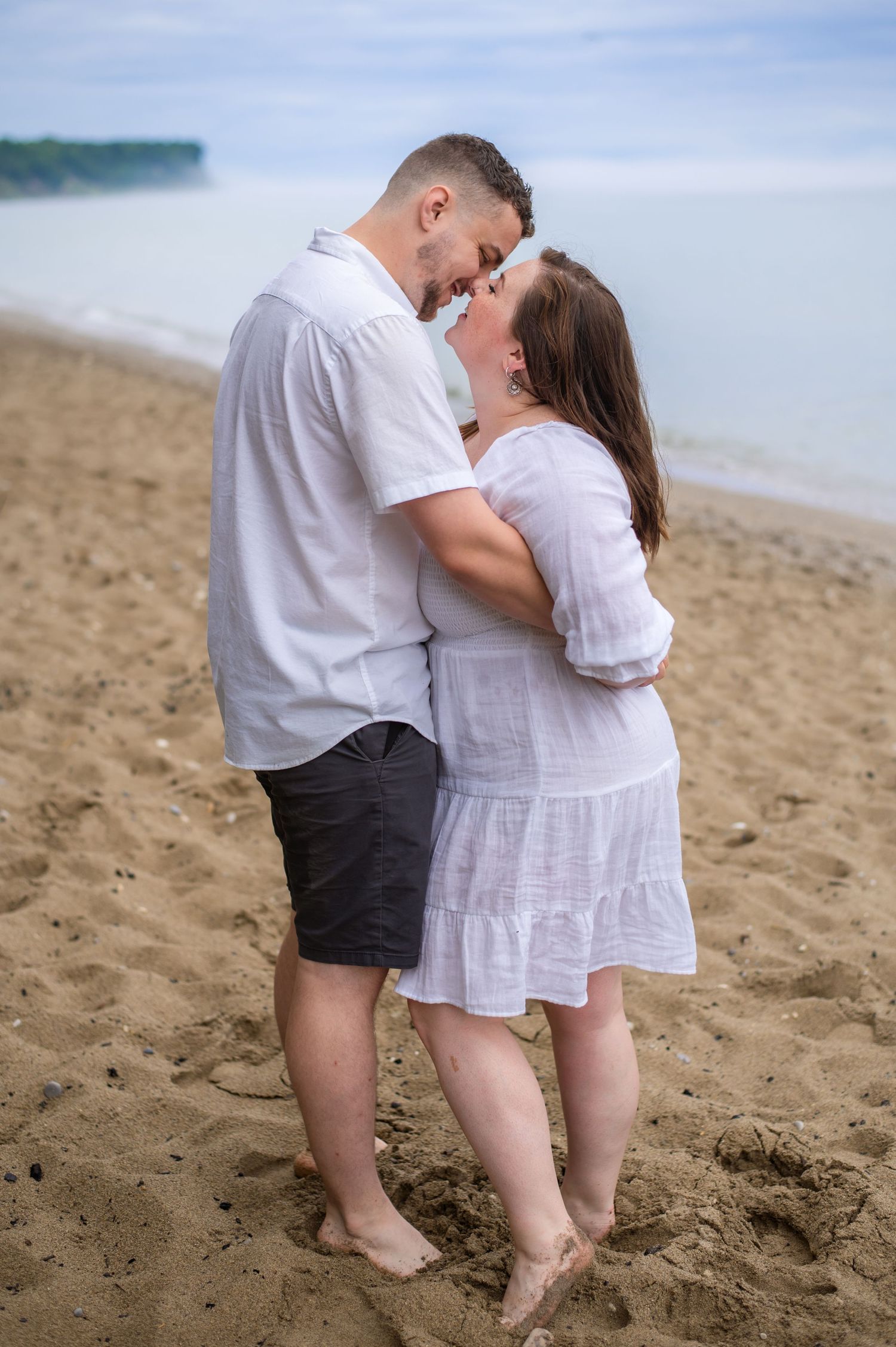 Romantic couple in white outfits embracing on sandy beach with water view.