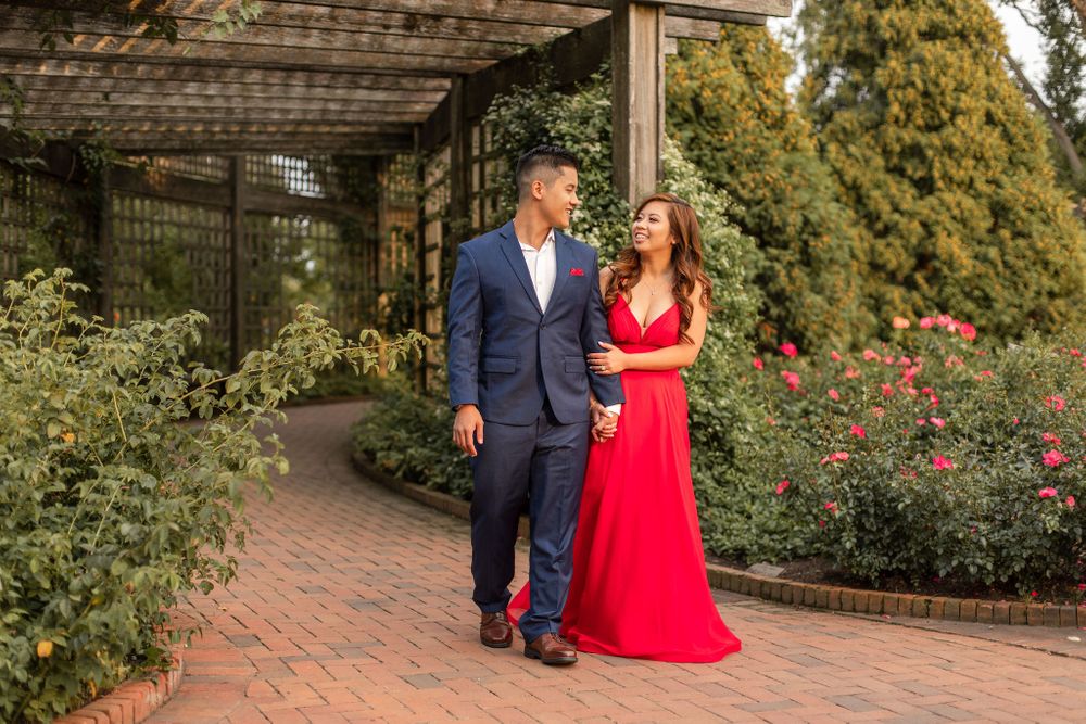 Couple poses in formal attire under garden pergola with blooming roses.