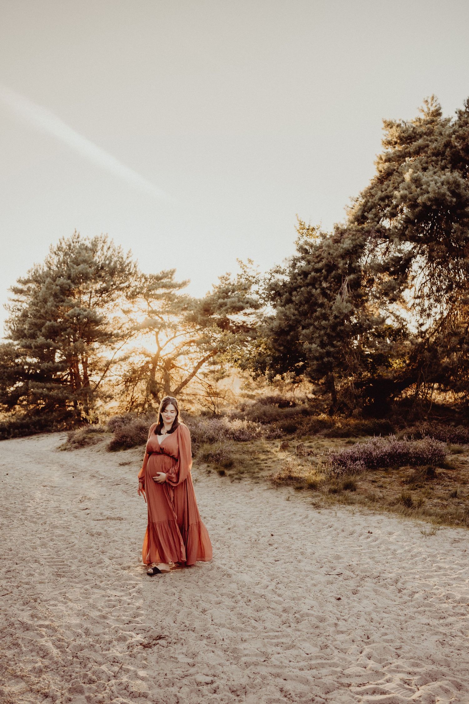 Figure in flowing rust-colored dress standing on sandy path with golden sunlit trees.