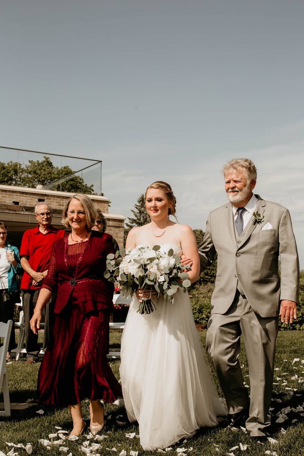 Bride walks down outdoor aisle with parents in gray suit and burgundy dress on sunny day.