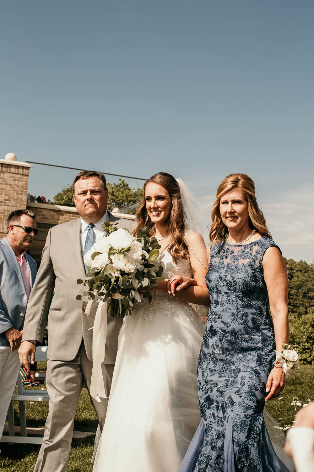 Smiling bride walks the aisle flanked by parents in gray suit and blue lace gown outdoors.