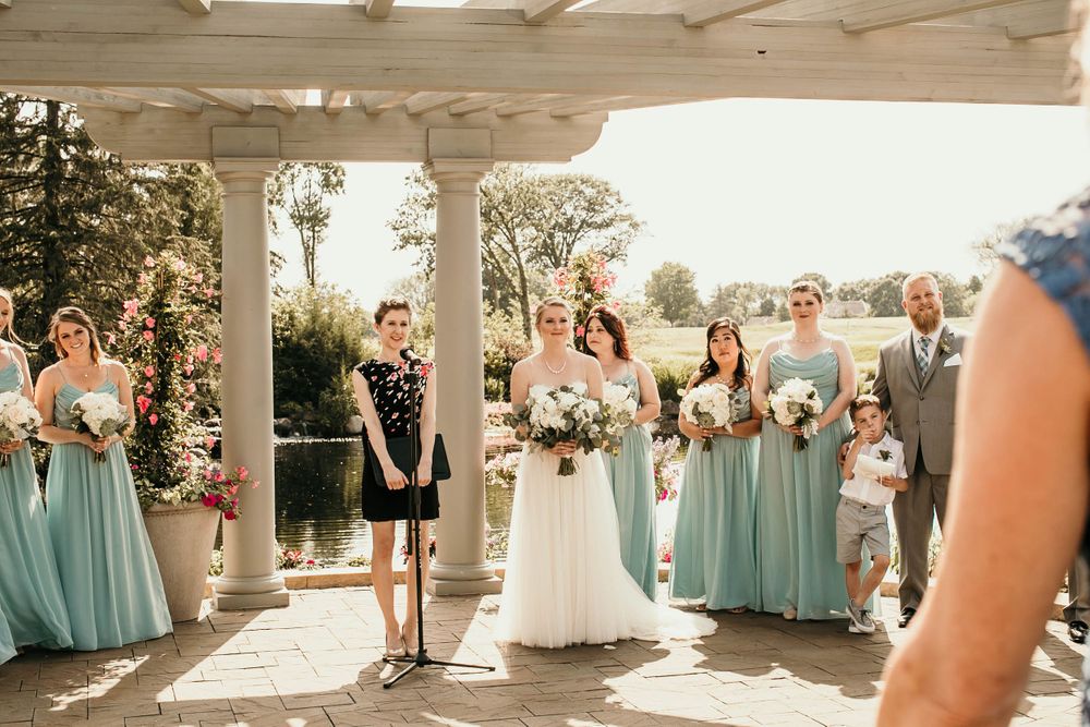 Bride in white gown stands with bridesmaids in mint dresses under pergola at outdoor ceremony.