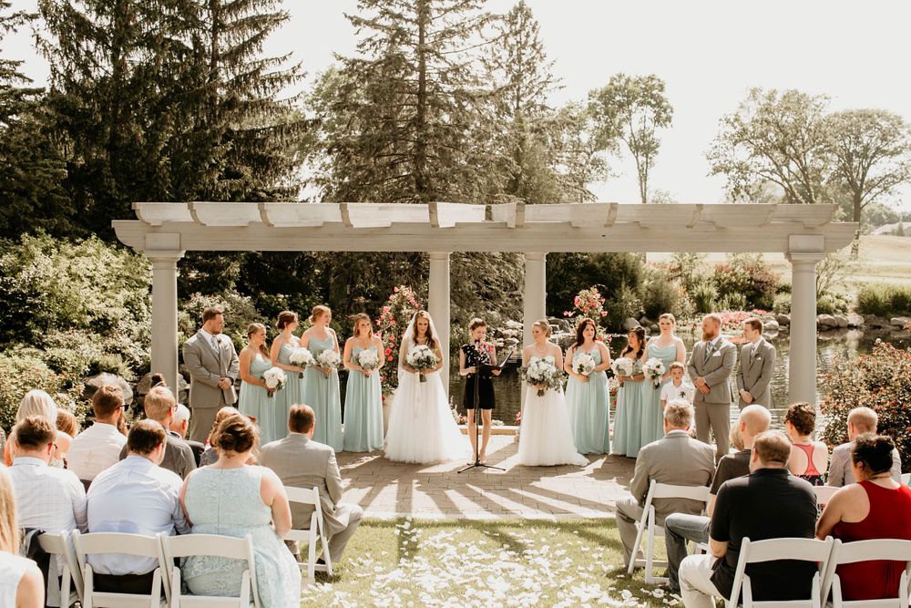 Outdoor wedding ceremony with bridesmaids in sage green dresses under a white pergola surrounded by lush greenery.