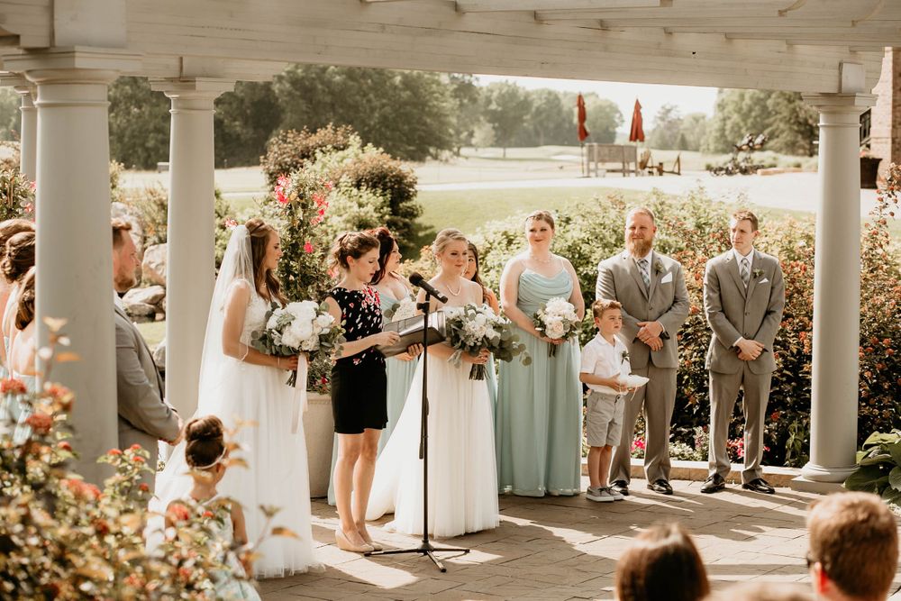 Two brides exchange vows on a flower-decorated porch with bridesmaids in mint dresses during outdoor ceremony.