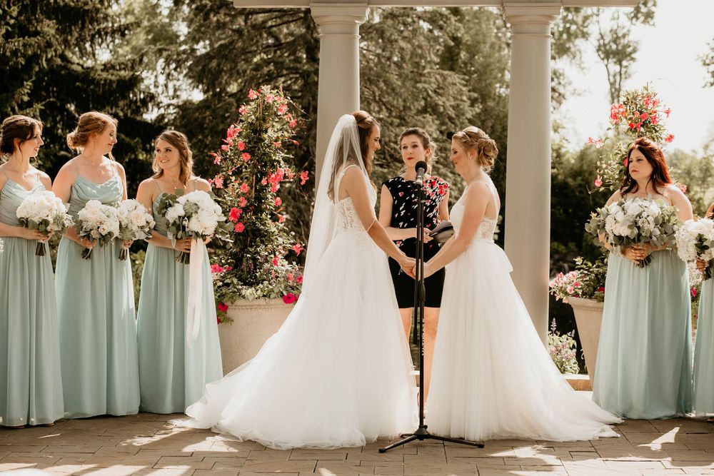 Two brides in white gowns hold hands at altar with bridesmaids in sage green dresses and pink floral backdrop.