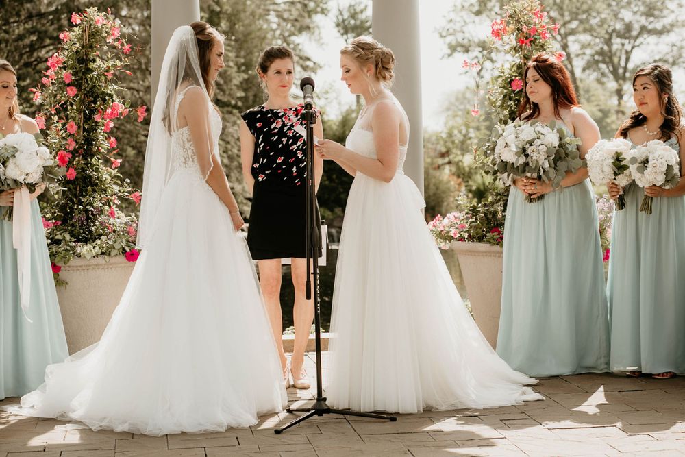 Two brides face each other at microphone during outdoor wedding vows with bridesmaids in mint green dresses.
