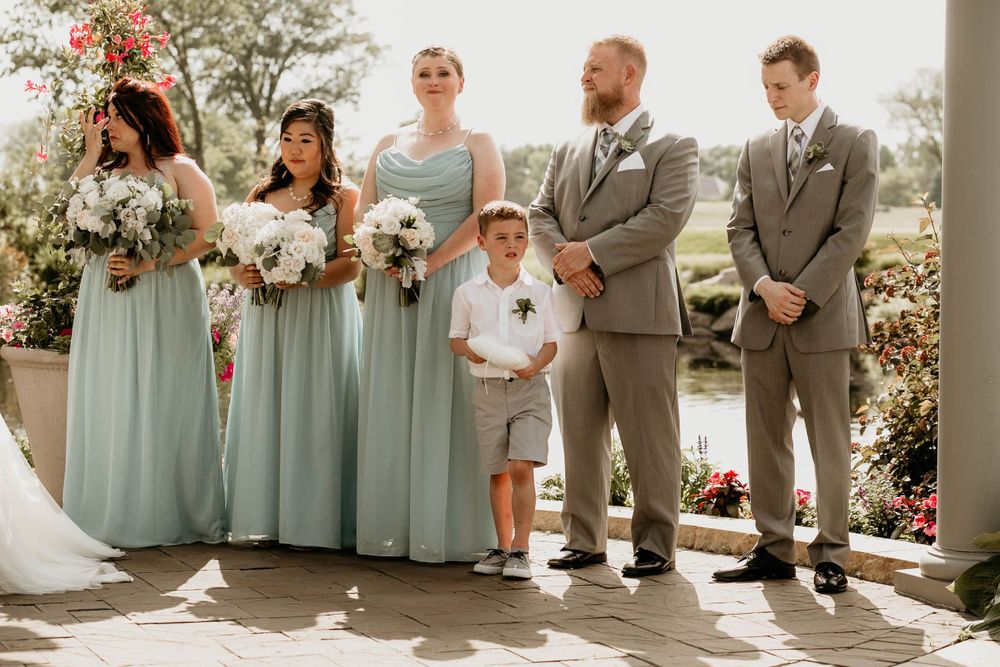 Wedding party members in sage green dresses and gray suits stand at outdoor ceremony with a young ring bearer.