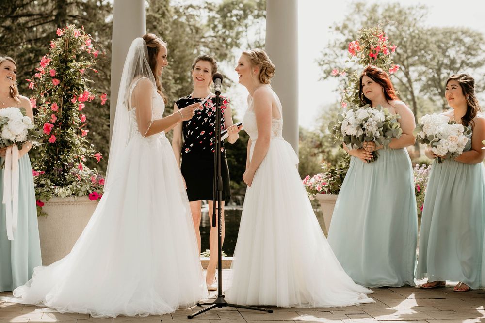 Two brides laugh joyfully during outdoor wedding ceremony with sage green bridesmaids and pink floral backdrop.