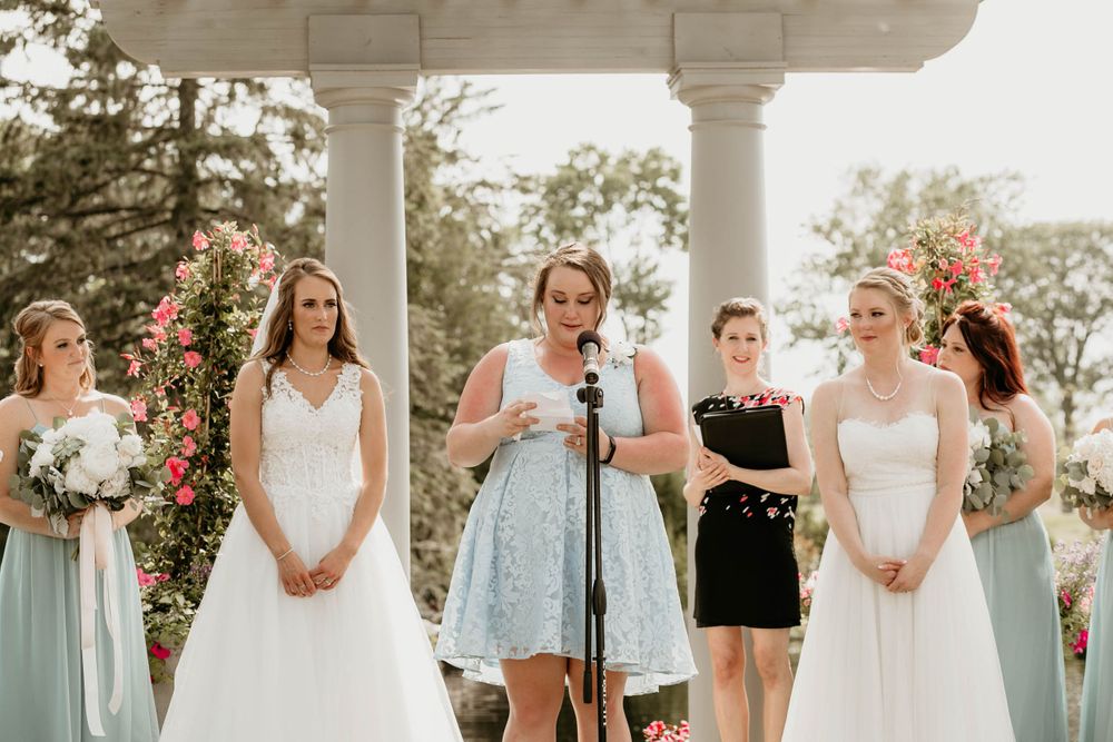 Wedding party member reads speech at microphone during outdoor ceremony with two brides and bridesmaids present.