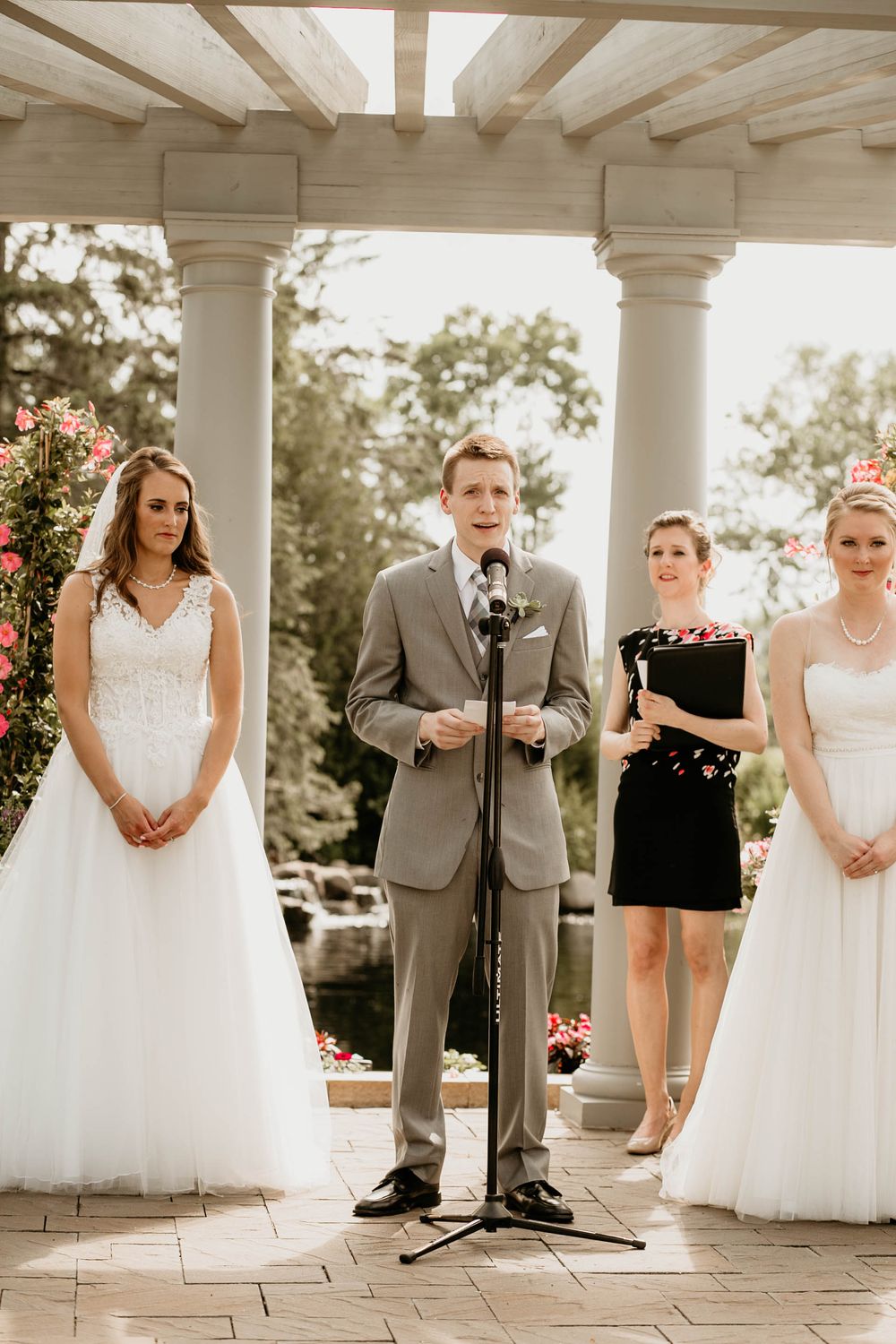 Groomsman speaks at microphone during outdoor wedding ceremony flanked by two brides and officiant under pergola.