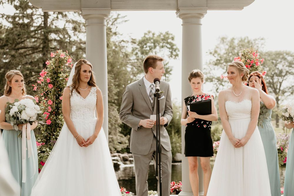 Two brides stand at outdoor wedding altar with bridesmaids in mint dresses and officiant at microphone.