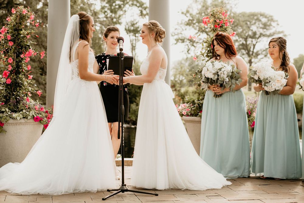 Two brides exchange rings at outdoor wedding ceremony with sage green bridesmaids and colorful floral backdrop.