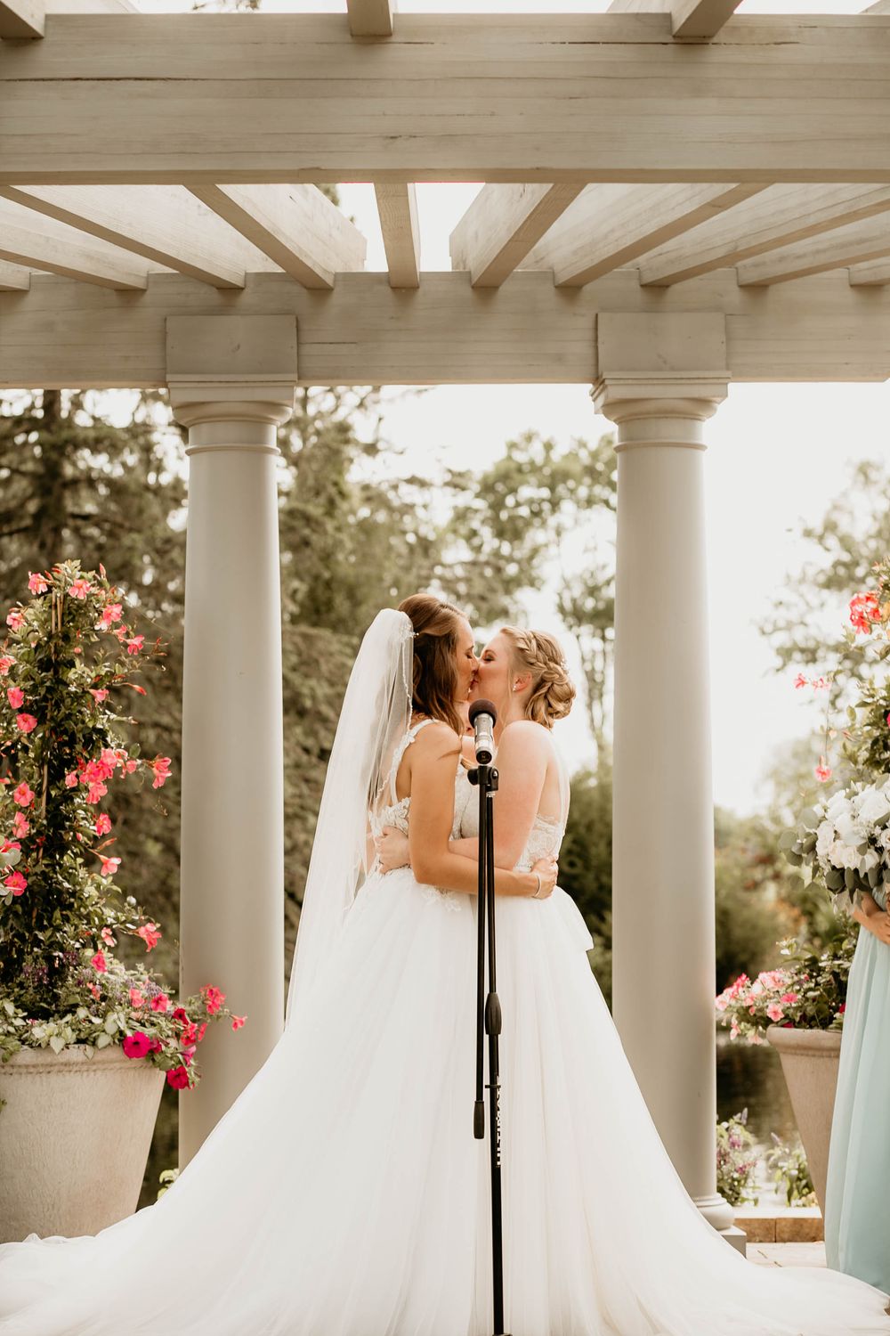 Two brides share first kiss under white pergola surrounded by pink roses at outdoor wedding ceremony.