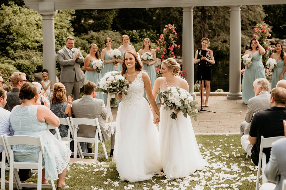 Two brides walk down flower petal aisle as guests applaud at elegant outdoor garden wedding ceremony.
