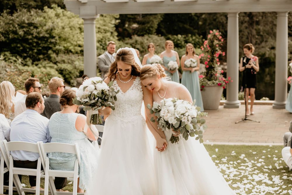 Two brides in white gowns hold bouquets and walk hand-in-hand down petal-lined aisle after ceremony.