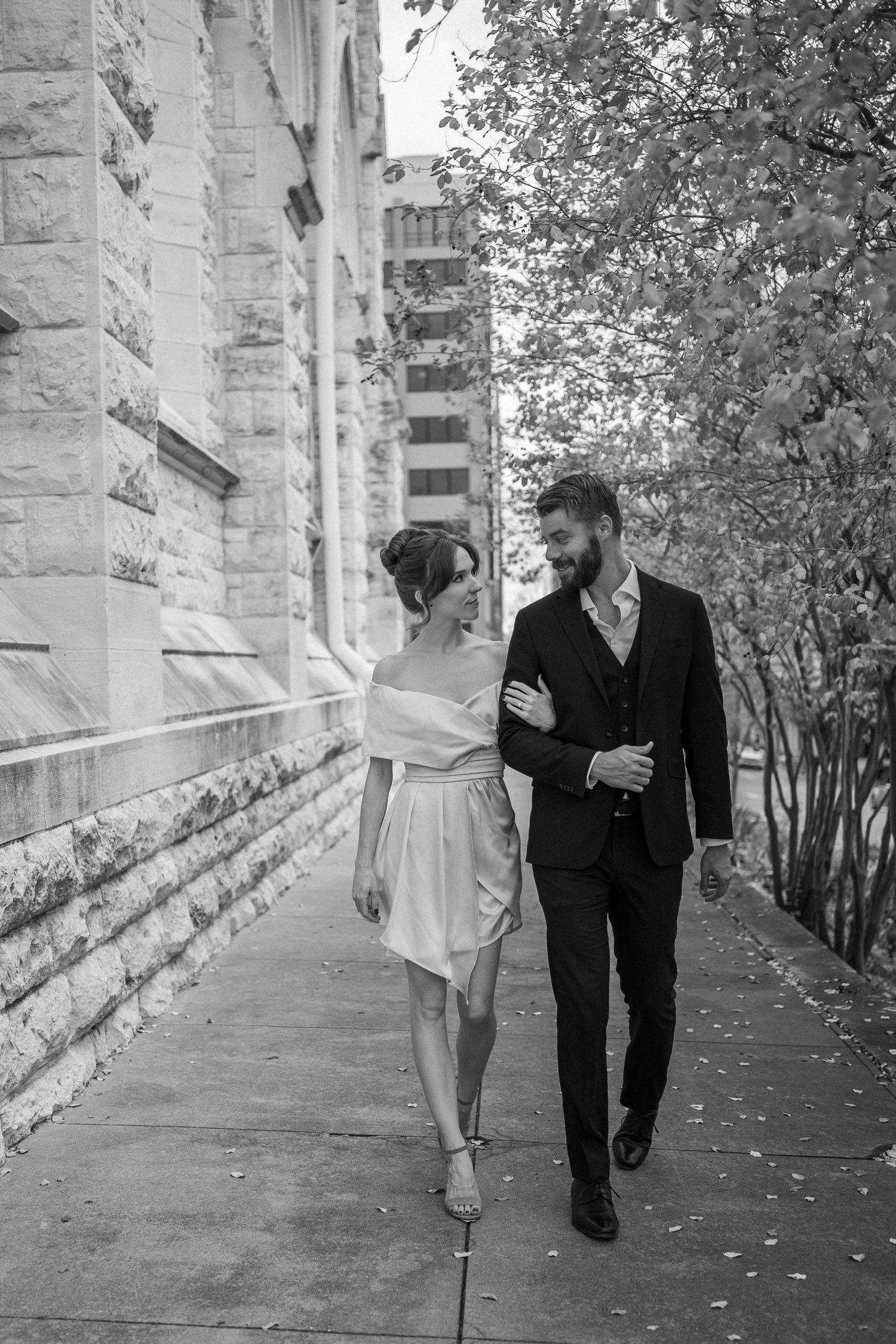 A couple walks arm in arm along a stone building pathway in formal attire.