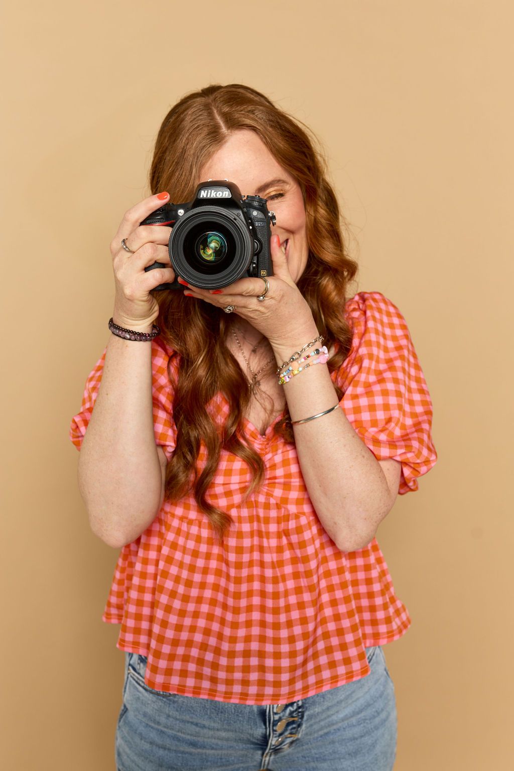 Photographer in orange gingham top taking picture with professional camera against beige background.