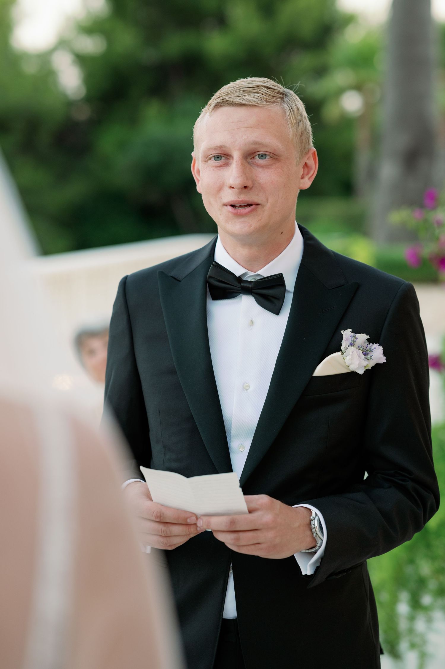 Groom in black tuxedo and bow tie reads from paper during outdoor ceremony.