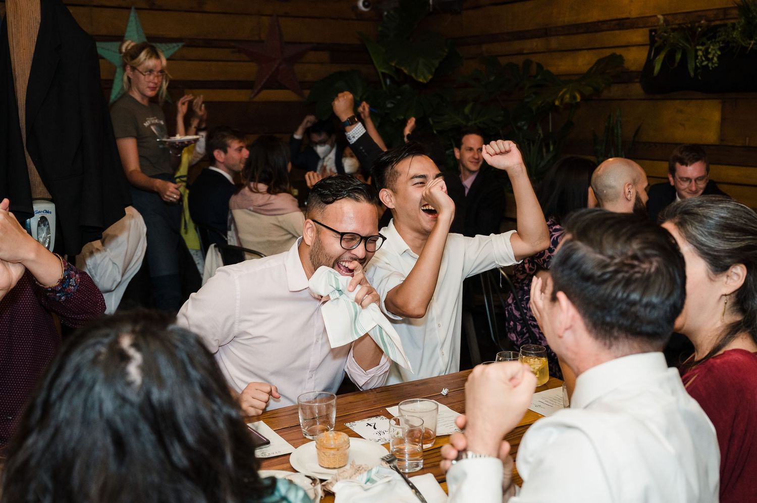 Guests celebrate energetically at a reception dinner with wooden walls and hanging plants.