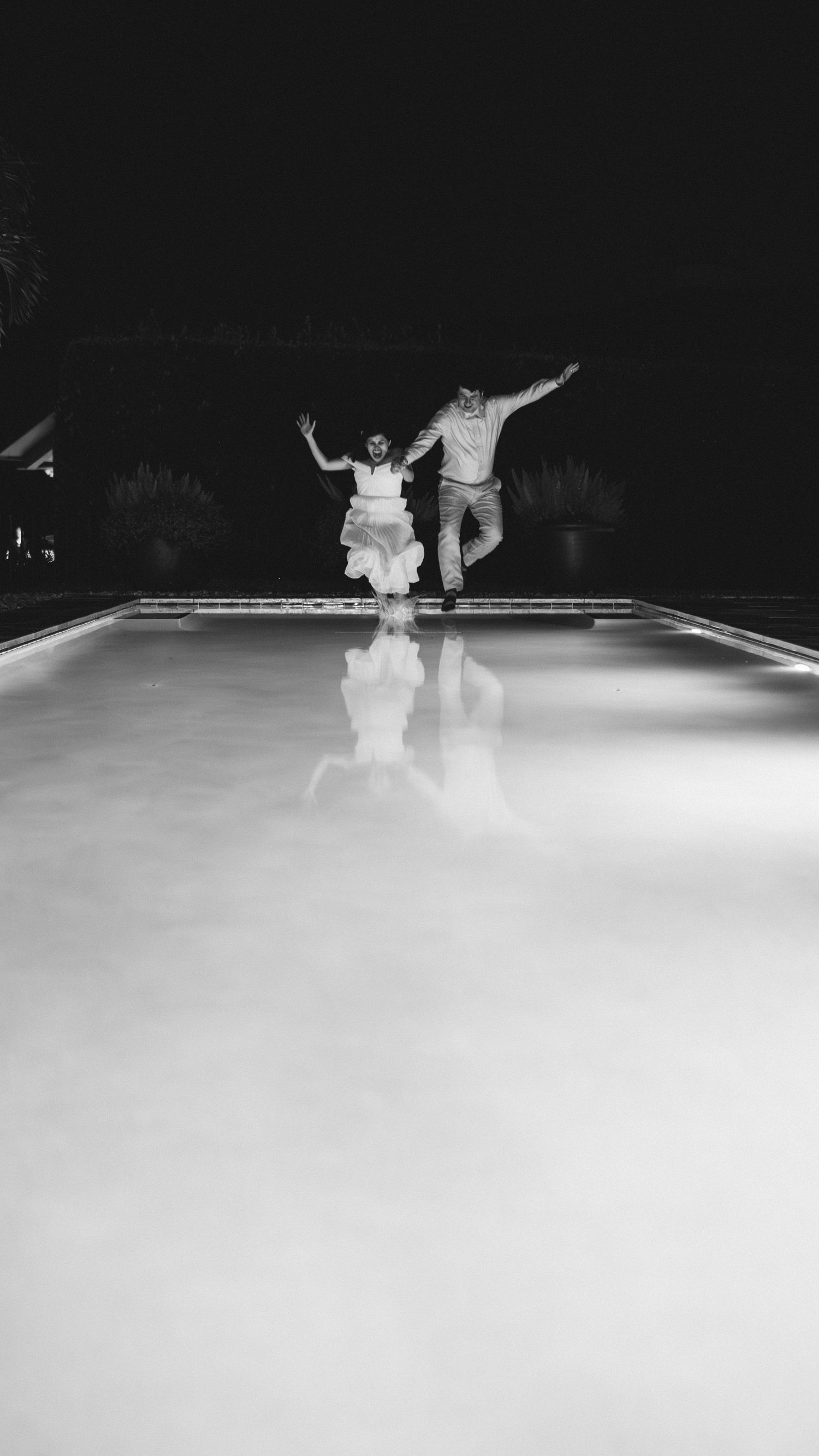 Silhouette of couple jumping together over reflective pool surface at night.