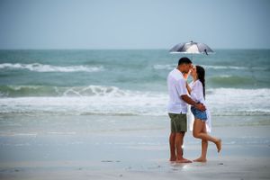 Couple embracing on beach with seagull flying overhead and ocean waves in background.