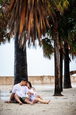 Romantic couple sitting under palm trees on tropical beach at sunset.